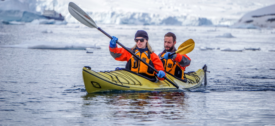 Two guests in their dry gear paddle a kayak in the frigid Antarctic waters during a sunny day.