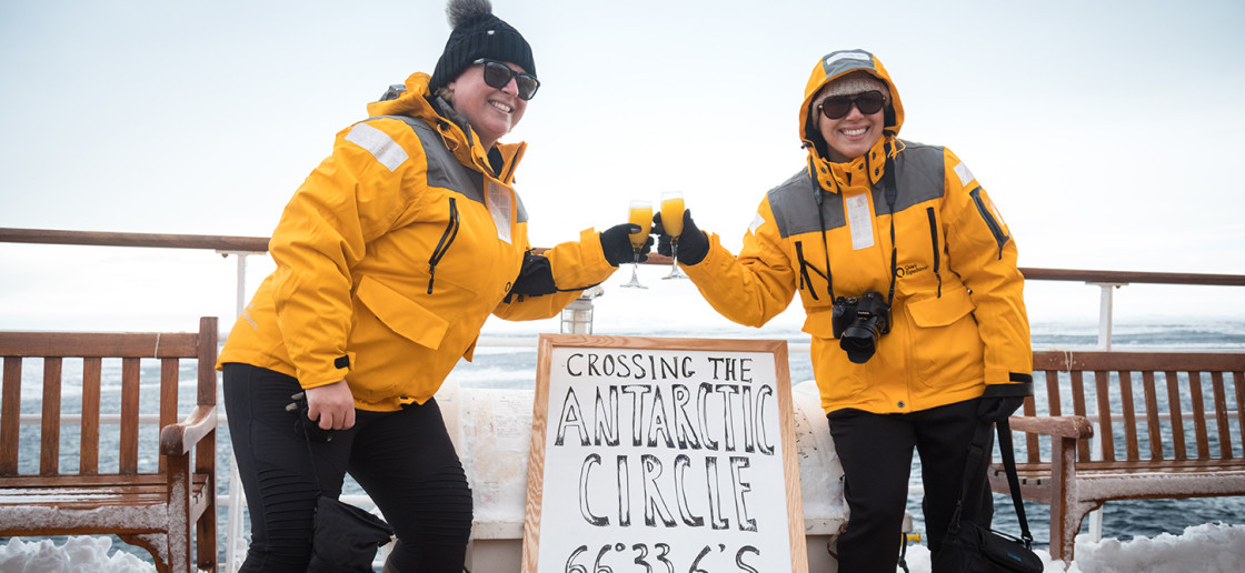 Two guests in yellow parkas pose for a photo next to a board that includes the coordinates of their ship, celebrating officially crossing the Antarctic Circle.