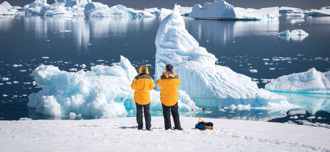 Two guests in yellow parkas admire and photograph rugged icebers off the waters of Port Charcot during a sunny afternoon in Antarctica.