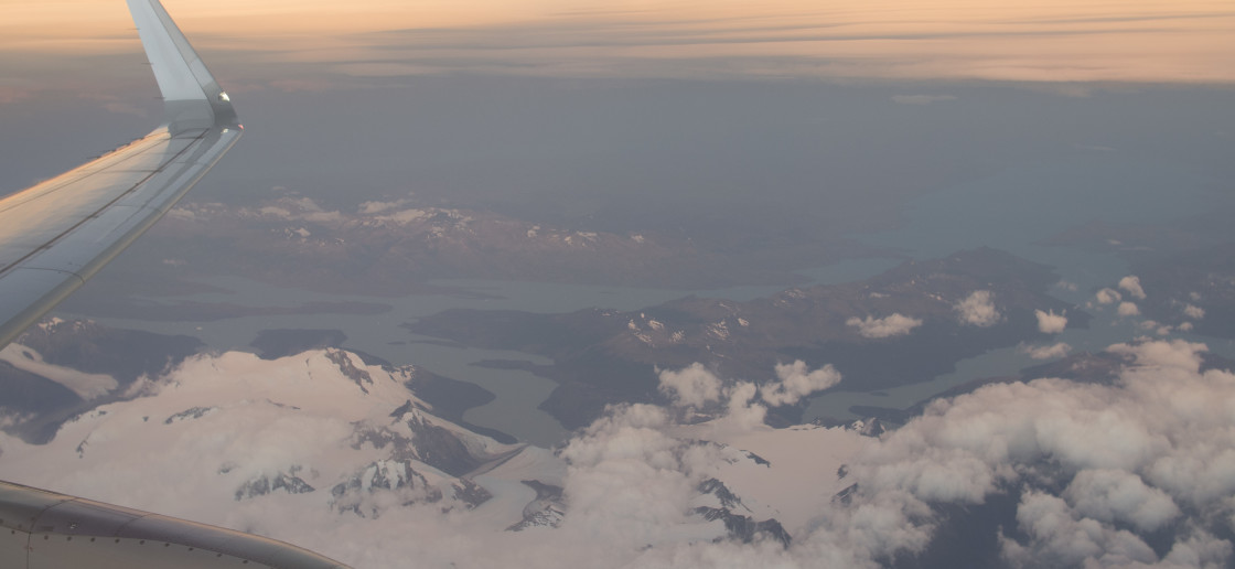 View from the window of an aircraft featuring its wing and a small cloud cover over the partially ice-covered peaks near Punta Arenas, Chile.