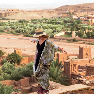 The girl-traveler in a hat is standing on a high wall with arms outstretched in the background of the city in the desert of Ait BenHaddou, Morocco, Africa