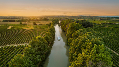A lone canal boat sails into the golden sunset on the Canal du Midi, cutting through vineyards and rural countryside for a picture-perfect moment on a French boating holiday.