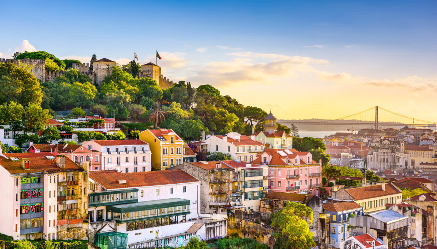 Bright colored buildings cover tree-filled hills along the Tagus River under a sunset sky in Lisbon, Portugal.