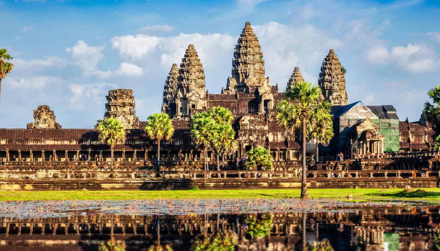 A view of Angkor Wat temple surrounded by palm trees and reflecting off a lake in Siem Reap, Cambodia.