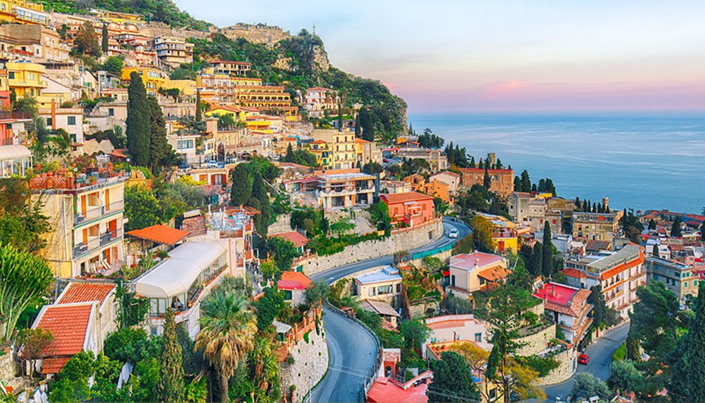 Bright, colorful houses line the coast of Taormina, Italy, in front of the Mediterranean Sea under a soft pink sunset.
