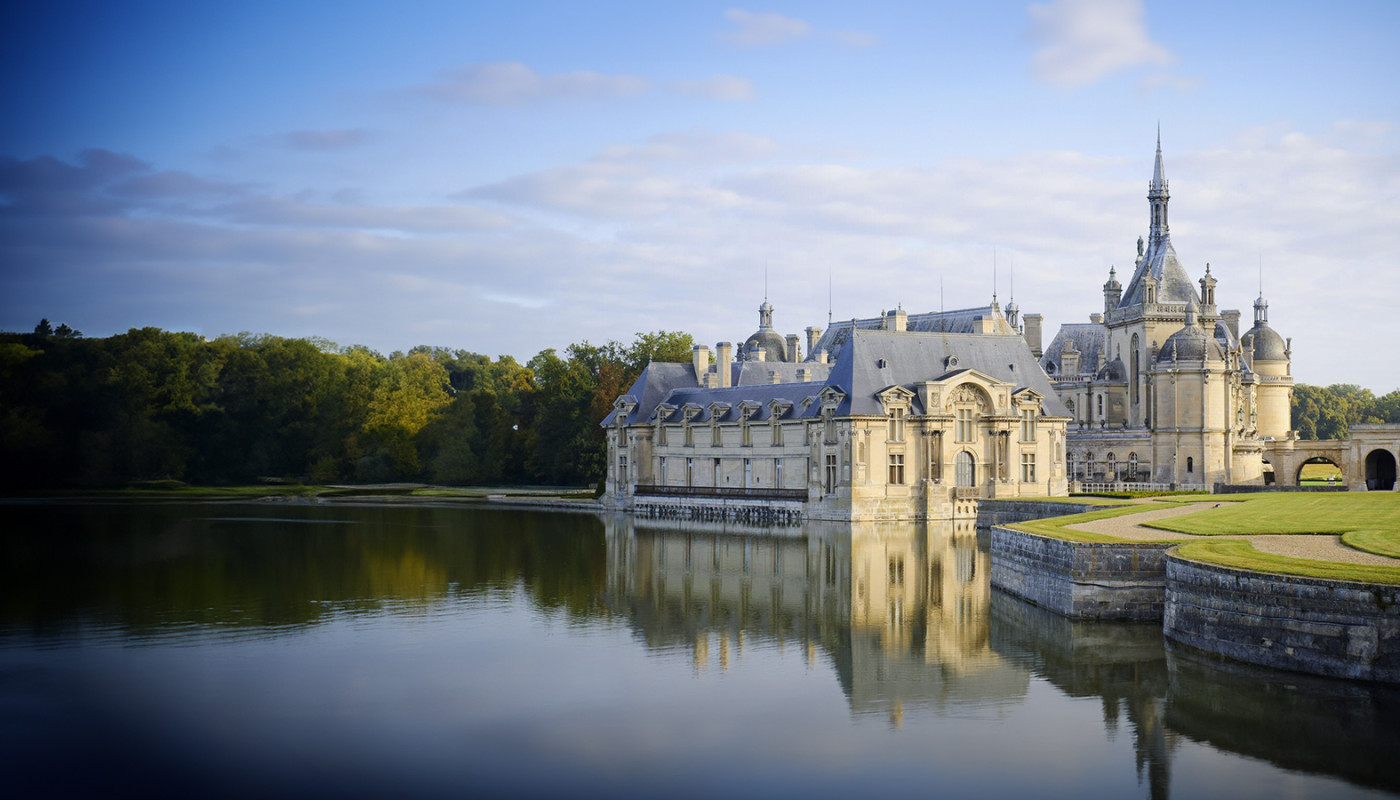 The Château de Chantilly reflects off the water at the edge of a moat under golden sunlight in Chantilly, France.