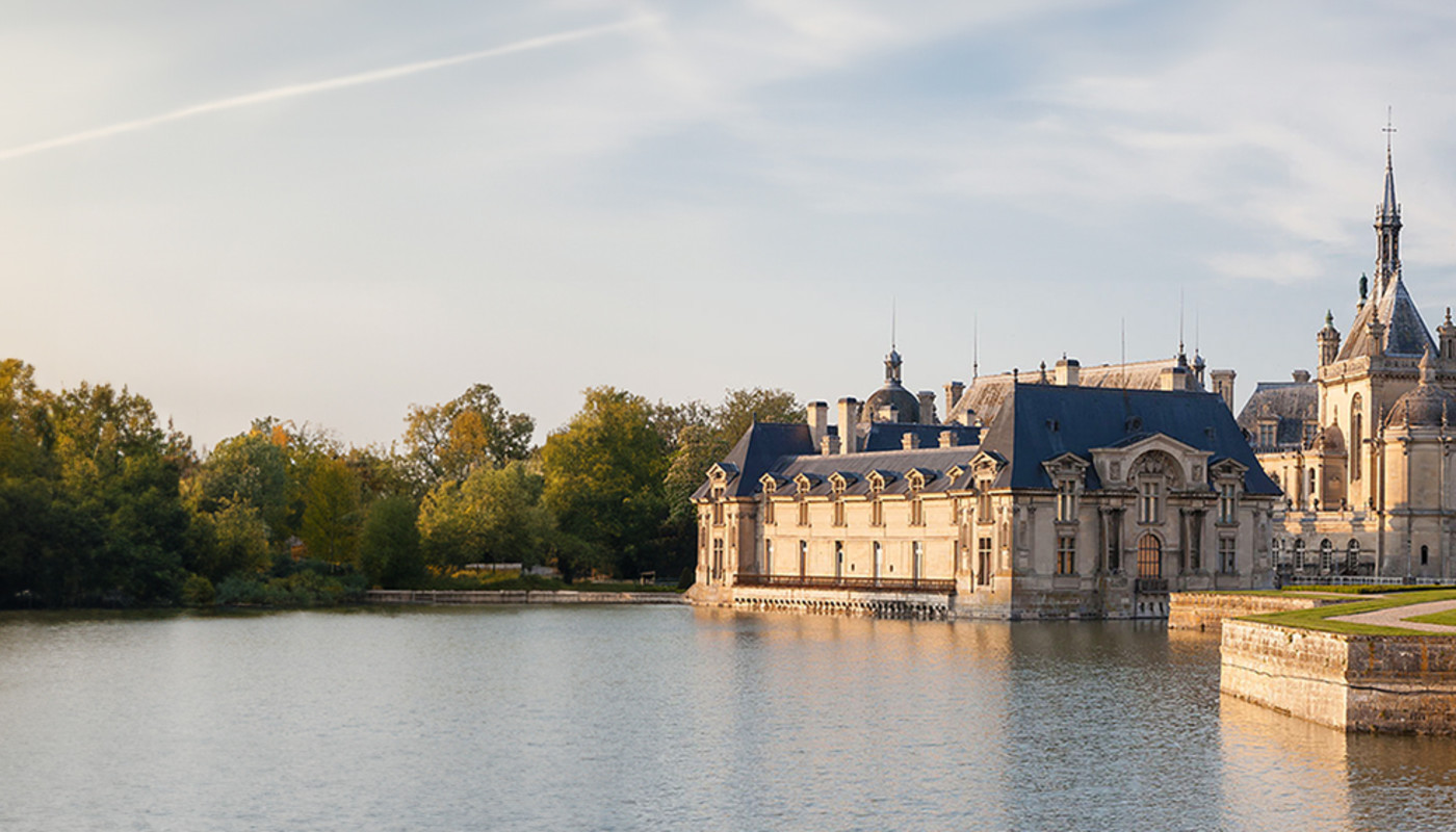 The Château de Chantilly reflects off the water at the edge of a moat under golden sunlight in Chantilly, France.