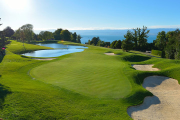 The green and bunkers on the 18th hole at Évian Resort Golf Club overlook Lake Geneva under blue sky in Évian-les-Bains, France.