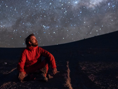 Junger Mann, auf dem Boden kniend, den Blick auf den Sternenhimmel gerichtet, in der Atacama-Wüste Chile