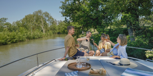 Group of friends toasting with wine on the bow of a houseboat under sunny skies.