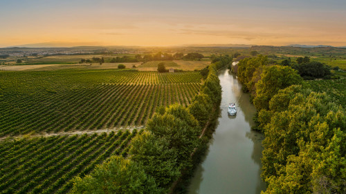 A lone canal boat sails into the golden sunset on the Canal du Midi, cutting through vineyards and rural countryside for a picture-perfect moment on a French boating vacation.