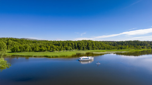Ein Stand-up-Paddler gleitet in der Nähe eines vertäuten Le Boat-Cruisers auf einem ruhigen See in Mecklenburg, umgeben von dichtem, grünem Wald.