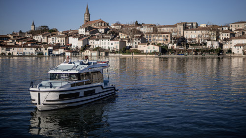 Un bateau Liberty naviguant paisiblement sur un bassin face à la ville de Castelnaudary, avec ses maisons et son clocher en arrière-plan sous un ciel dégagé.
