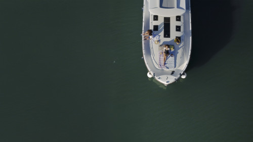 Familie entspannt sich auf dem Sonnendeck des Boots; eine Person angelt, während eine andere auf einer Decke über dem ruhigen, grün schimmernden  Wasser an Deck liegt.