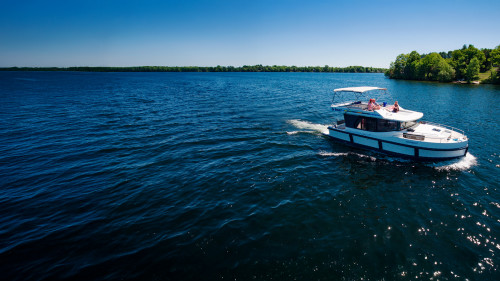 Aerial view of a Le Boat cruiser gliding along the calm Rideau Canal in Canada, surrounded by lush green forests under a clear blue sky.