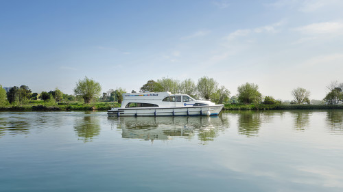 Comfort category boat navigating along a peaceful river bordered by lush trees and open landscape.