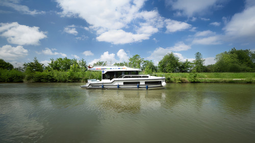 Premier category boat cruising on a calm river under a clear blue sky, surrounded by green countryside.