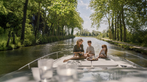Mum and kids enjoying a peaceful breakfast together on the bow of the boat along a tree-lined canal.