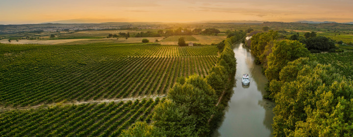 A lone canal boat sails into the golden sunset on the Canal du Midi, cutting through vineyards and rural countryside for a picture-perfect moment on a French boating holiday.