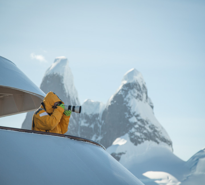 A guest in a yellow parka stands on one of the top decks of their vessel with a telephoto lens. Mountain peaks can be seen in the distance.