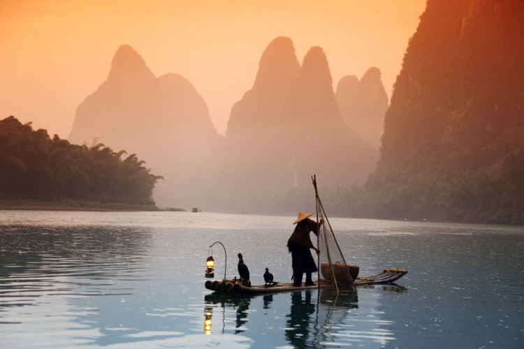 Silhouette eines Fischers und Kormoranen auf dem Li-Fluss in Yangshuo, China, im Hintergrund das Karstgebirge im Licht des Sonnenuntergangs