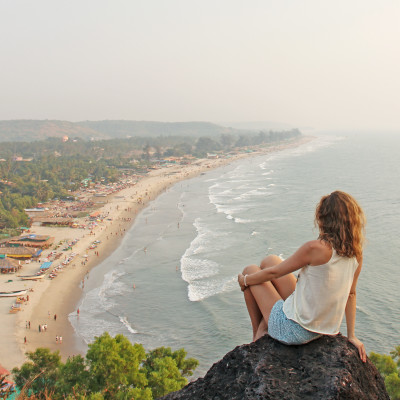 A young girl with blond hair sits on top of a mountain and looks at the sea and beach, Arambol, Goa, India