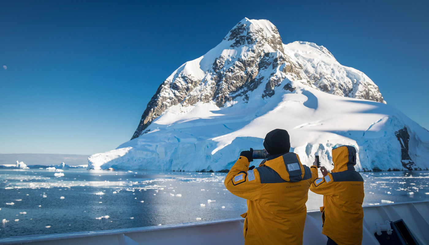 Two guests in yellow parkas take photos of snow-covered mountains while the ship passes through the Lemaire Channel.