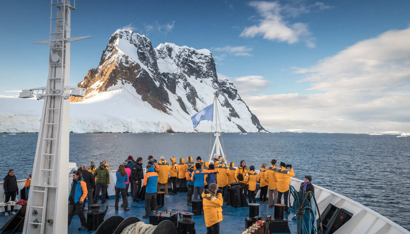 Passengers stand on the bow of the ship, as the peaks of the Lemaire Channel tower over them