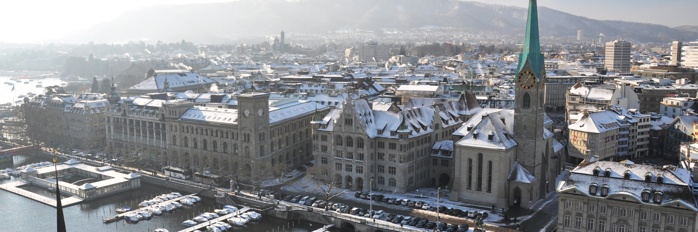 Aerial winter view of Zurich, Switzerland, with snow-covered rooftops across the historic city center. The Limmat River runs through the foreground, lined with snow-dusted boats at docks. The prominent Fraumünster Church with its distinctive green spire dominates the right side, while the large Zurich City Hall (Rathaus) stands along the riverbank. Snow-capped hills and mountains are visible in the background under a hazy winter sky.