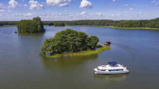 Ein Le Boat-Hausboot fährt an kleinen grünen Inseln auf einem weiten See in Deutschland vorbei, unter einem leuchtend blauen Himmel mit fluffigen Wolken.