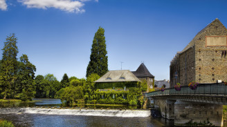 Picturesque riverside scene in Malestroit, Brittany, with a stone mill, ivy-covered house, and lush green trees under a clear blue sky.