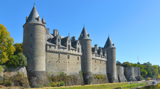 The medieval Josselin Castle, with its conical turrets, rises above the Oust River, its stone walls mirrored in the calm water and framed by lush trees under a clear blue sky.