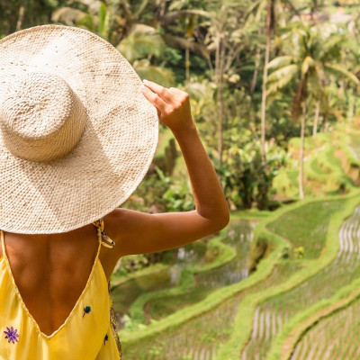 Is Indonesia safe? Beautiful young lady in shine through dress touch straw hat. Girl walk at typical Asian hillside with rice farming, mountain shape green cascade rice field terraces paddies. Ubud, Bali, Indonesia