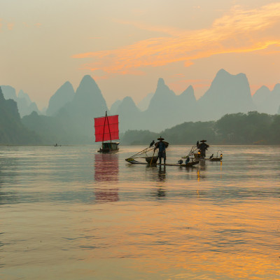 Fisherman stands on traditional bamboo boats at sunrise (boat with a red sail in the background)