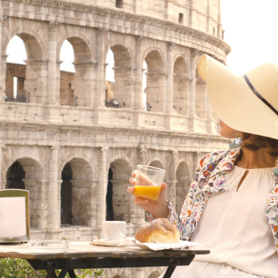 Happy young woman tourist drinking coffee and juice with cornetto at the table outside a bar restaurant in front of the Colosseum in Rome