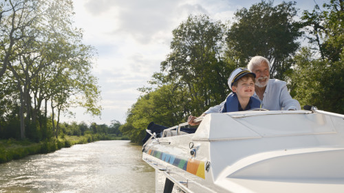 Grandfather and grandson steering a canal boat together on a sunny countryside journey.