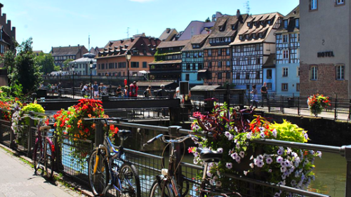 Traditionelle Häuser im Straßburger Stadtteil „Petite France“ unter blauem Himmel, entlang des von bunten Blumen gesäumten Kanals.