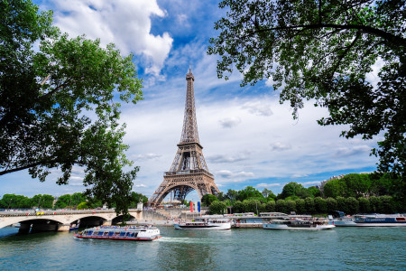 Eiffel Tower rising above the Seine River, framed by green trees, with tour boats and a bridge in Paris, France.