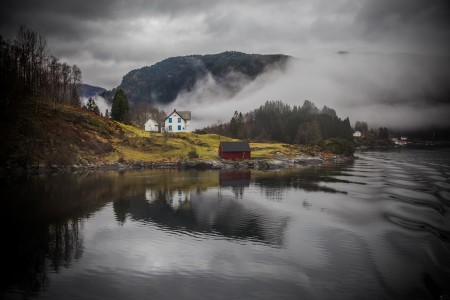 White house and red boathouse on a misty Norwegian fjord shoreline near Bergen, with fog-draped mountains reflected in calm water.