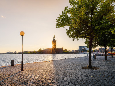 Stockholm City Hall at sunset, viewed from a cobblestone waterfront promenade with a lamp post and tree in the foreground.