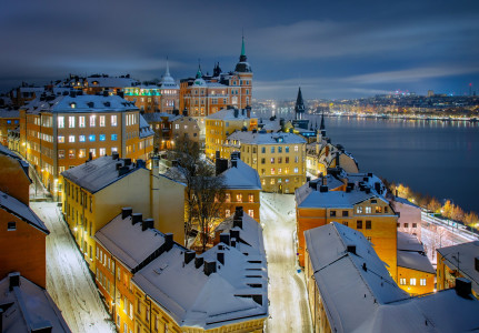 Aerial night view of Södermalm district, Stockholm, with snow-covered rooftops, glowing streets, and waterfront city lights.