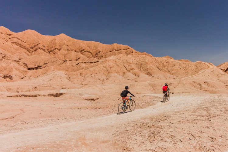 Zwei Radfahrer auf einem Sandweg in der Atacama-Wüste, Chile, mit roten Felsen und blauem Himmel.