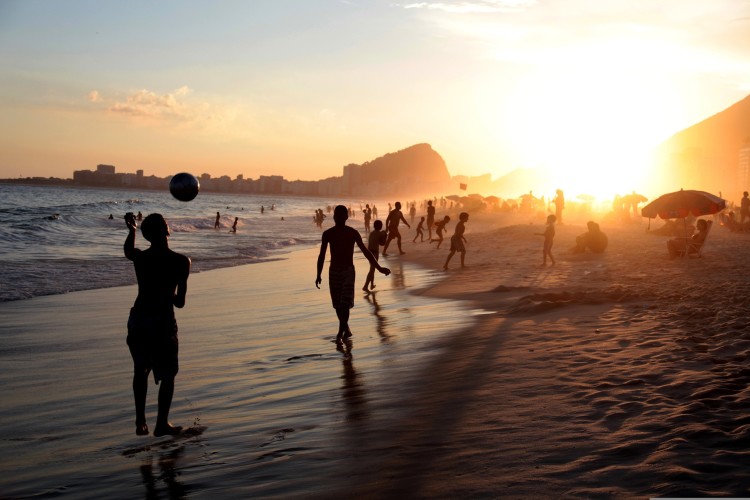 Menschen spielen Fußball am Strand von Rio de Janeiro bei Sonnenuntergang, Silhouetten vor goldenem Himmel.