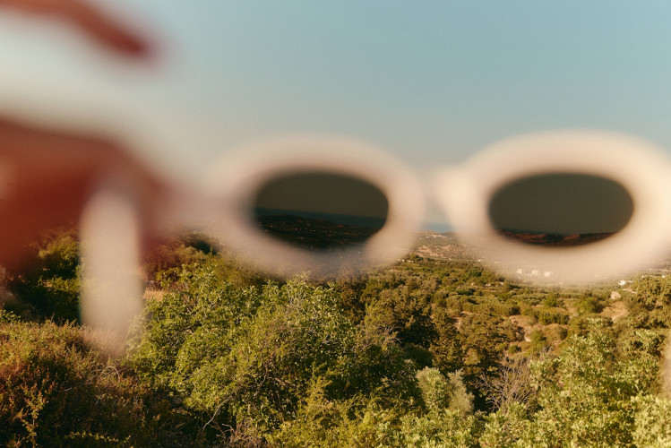 Hand holding white oval sunglasses up to a lush green hillside landscape under a blue summer sky.