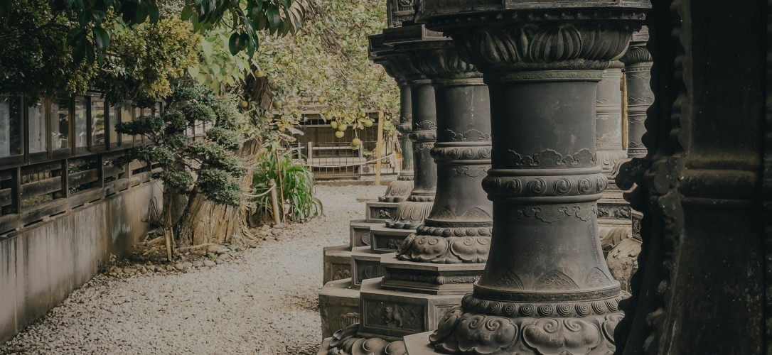 Verzierte Steinsäulen eines japanischen Tempels neben einem Kiesgarten mit Bäumen und traditionellem Holzgebäude.