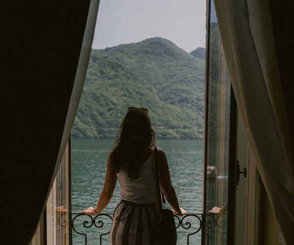 Woman standing at open balcony doors, gazing at a serene lake and lush green hills beyond.