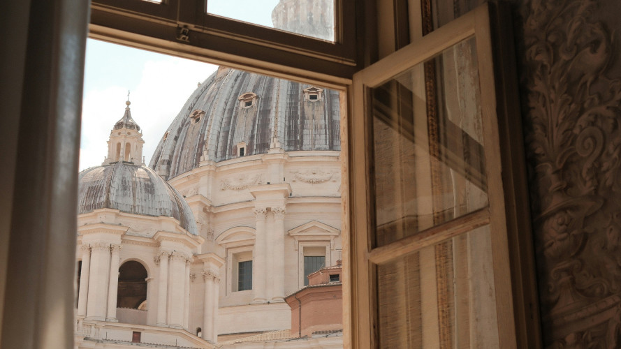 View through an open wooden window of St. Peter's Basilica's dome in Vatican City, with ornate carved wall detail visible.