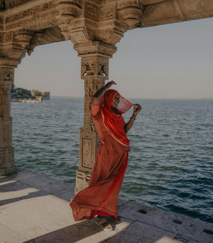 Woman in red saree posing beside ornate carved stone columns of an Indian temple overlooking a lake.
