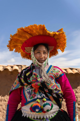 Peruvian woman in traditional colorful embroidered clothing and a large orange fringed hat, standing outdoors under a blue sky.