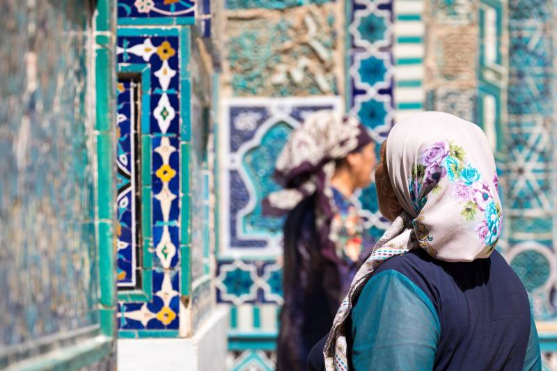 Verschwommen im Vordergrund zwei traditionell gekleidete Frauen vor dem Hintergrund des historischen Friedhofs Shahi Zinda in Samarkand, Uzbekistan